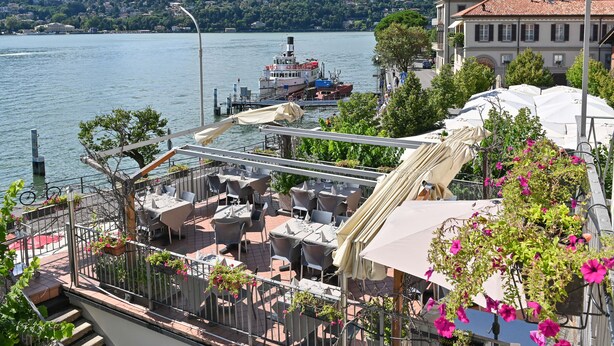A riverside restaurant with outdoor seating, a dock with a boat, and a mountainous backdrop. - Hotel Marco's (Como)