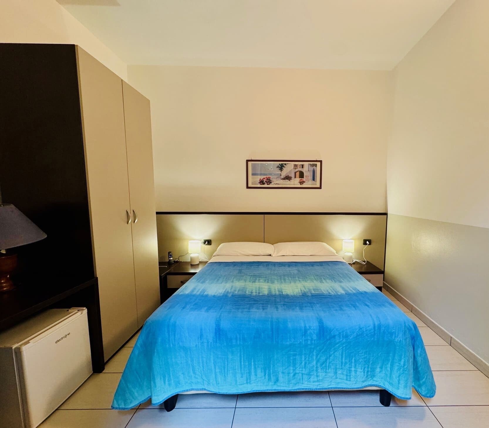 Quarto de hotel com cama, mesa de cabeceira, luminária, mini-geladeira e um quadro na parede.