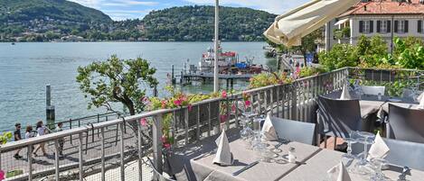 A lakeside restaurant with tables set for dining, overlooking a lake with boats and a hill in the background.