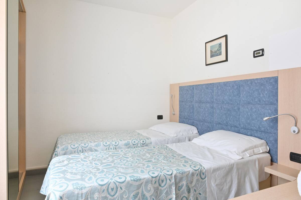 A room with two beds, a bedside table, a wall-mounted light, and a small picture frame.