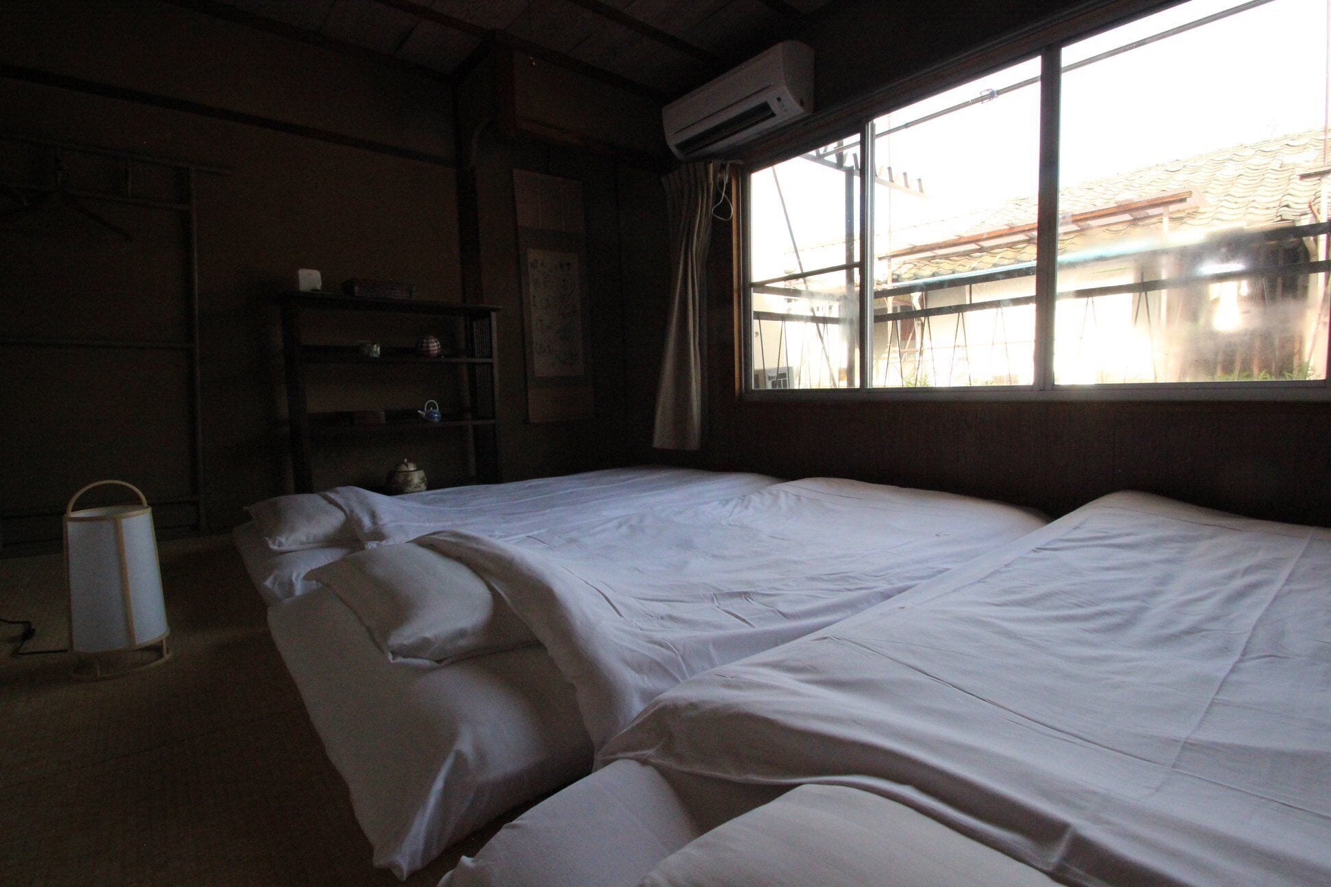 A room with two beds, a shelf with books and a kettle, and a window with curtains.