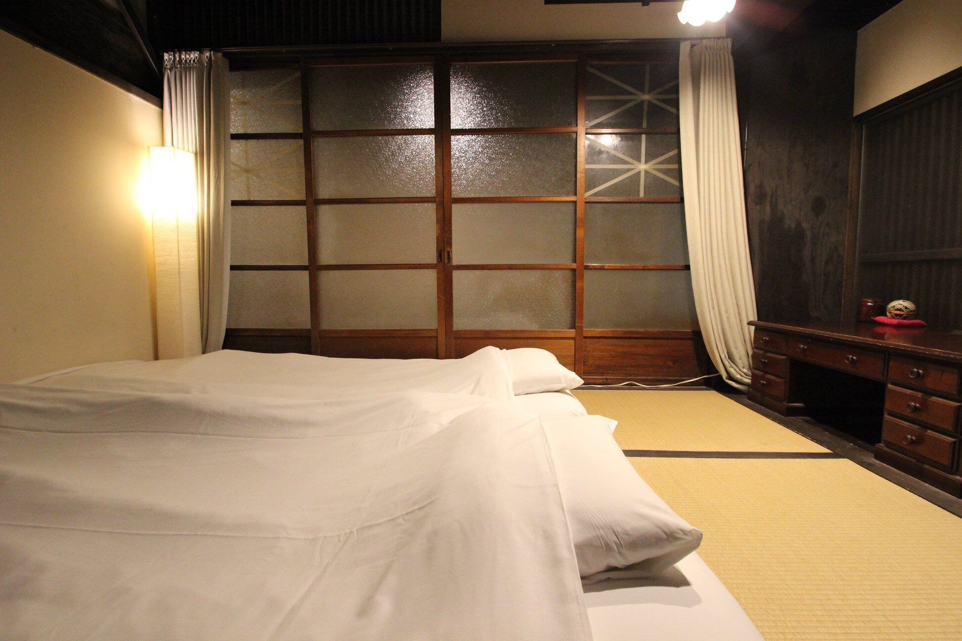 A traditional Japanese-style room with tatami flooring, a low wooden bed, sliding shoji doors, and a dark wooden dresser.