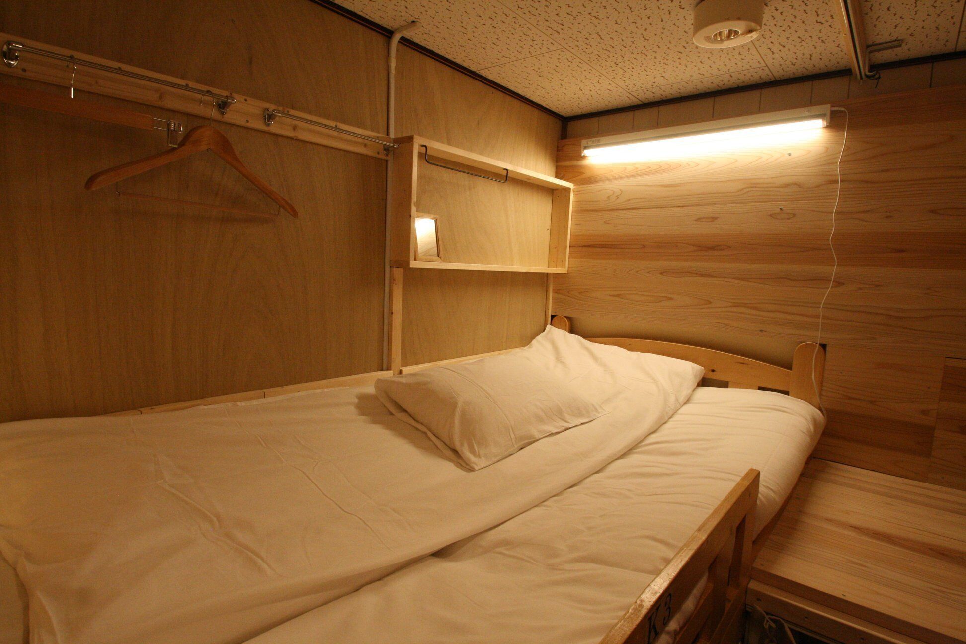 A small, wooden room with a single bed, a hanging coat rack, and a shelf with a light above it.