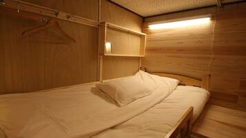 A small, wooden room with a single bed, a hanging coat rack, and a shelf with a light above it.