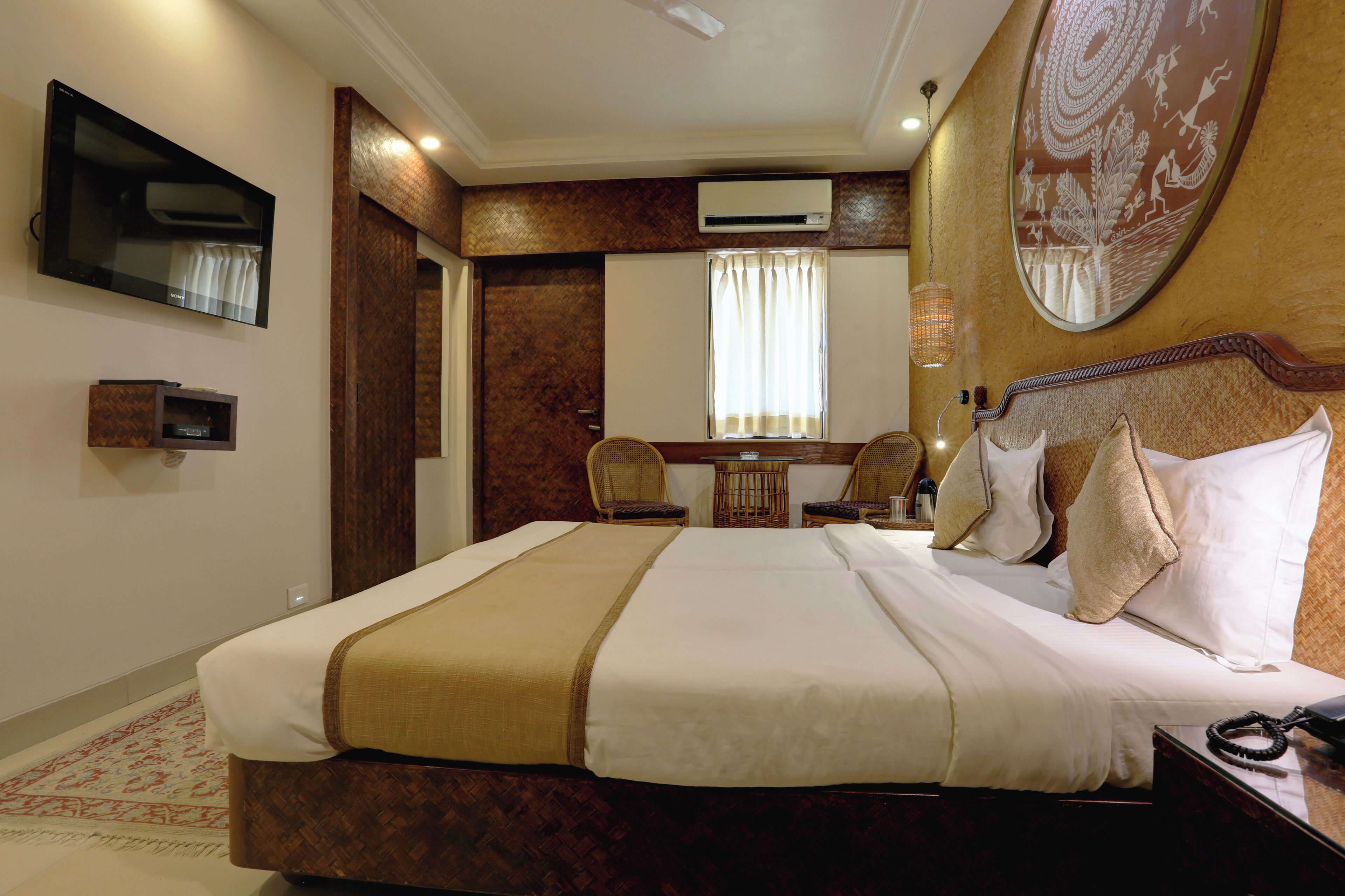 Habitación de hotel con una cama grande, un escritorio con sillas, un televisor de pantalla plana, un equipo de aire acondicionado y un espejo decorativo.