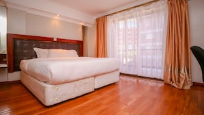 A bedroom with a large bed, wooden flooring, and a window with sheer curtains. - Nairobi Upperhill Hotel (Nairobi)