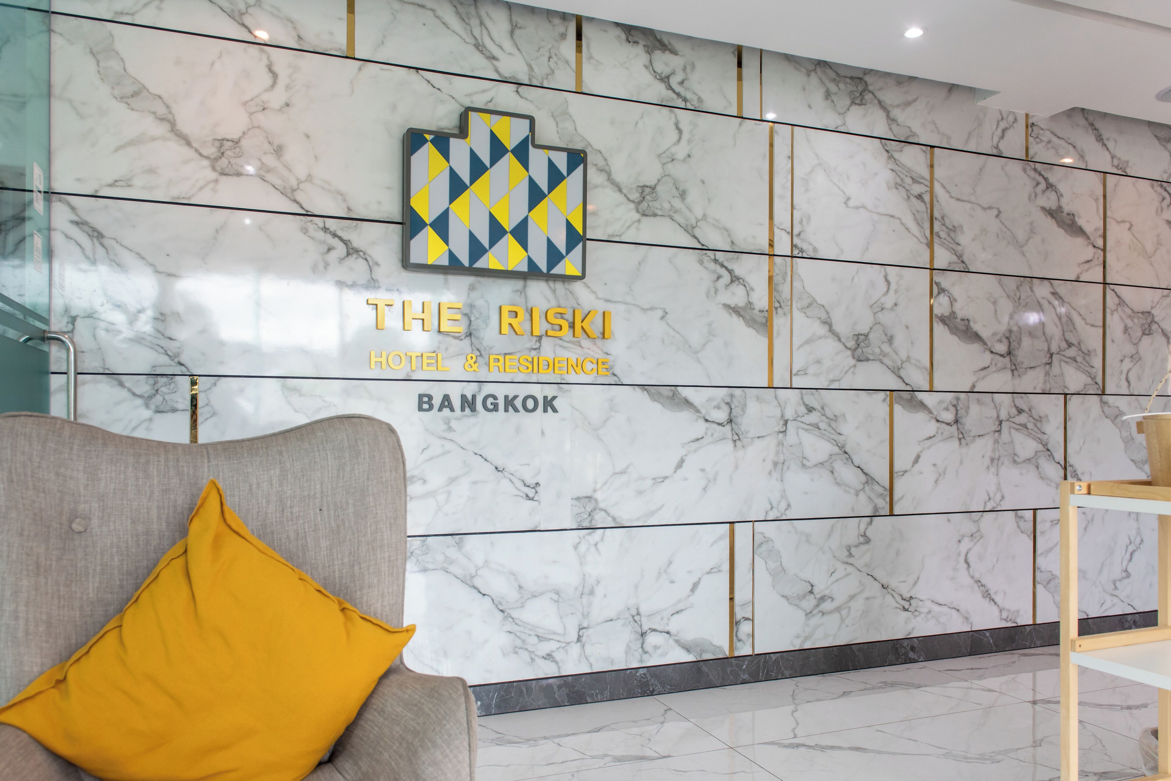 A reception area with a marble wall, a logo, and the text "THE RISKI HOTEL & RESIDENCE BANGKOK".