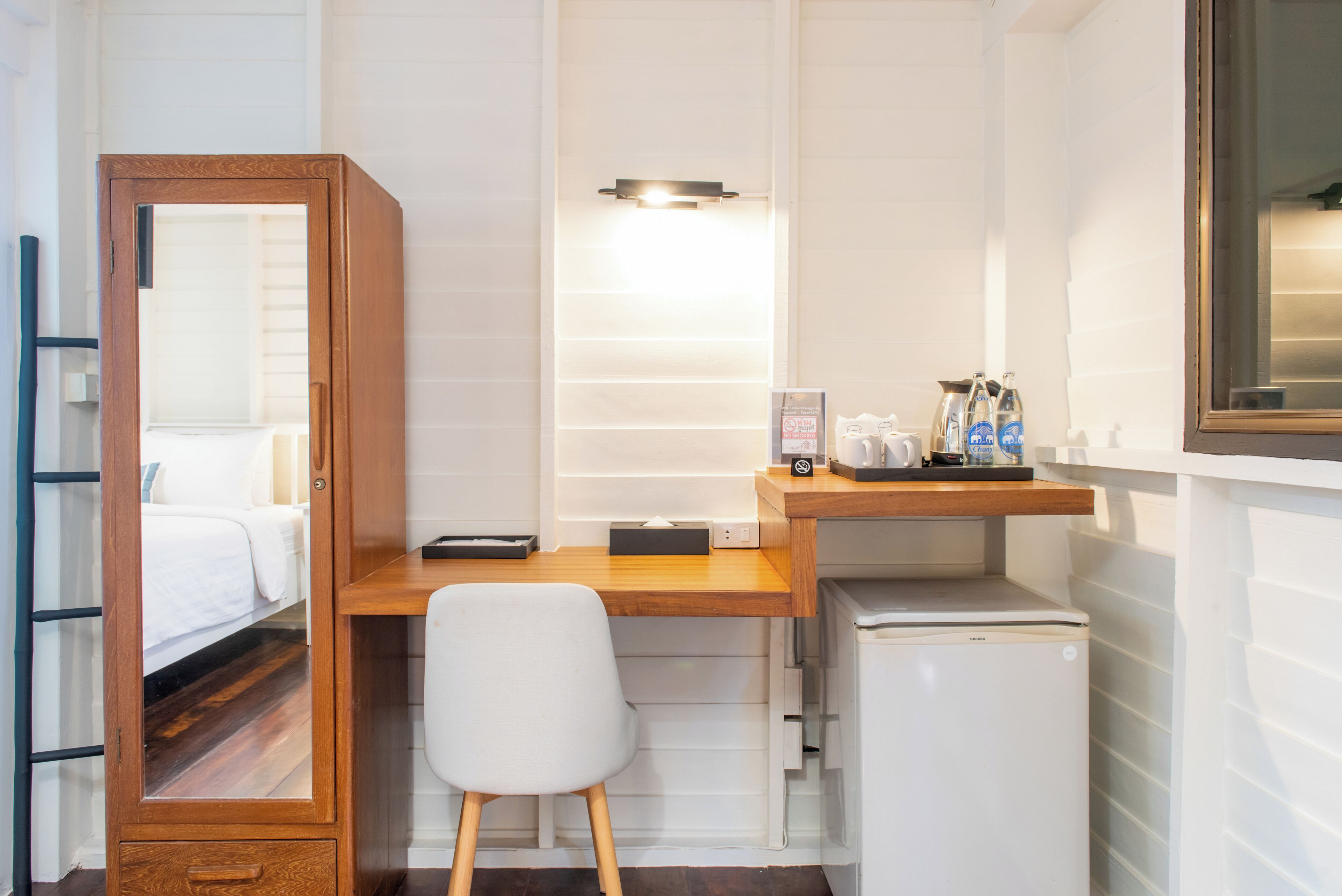 A compact room with a wooden desk, a white chair, a small refrigerator, and a bed with white bedding.