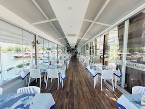 A restaurant with glass tables and chairs, wooden flooring, and large windows offering a view of boats on water. - Berg Hotel (Ortaca)