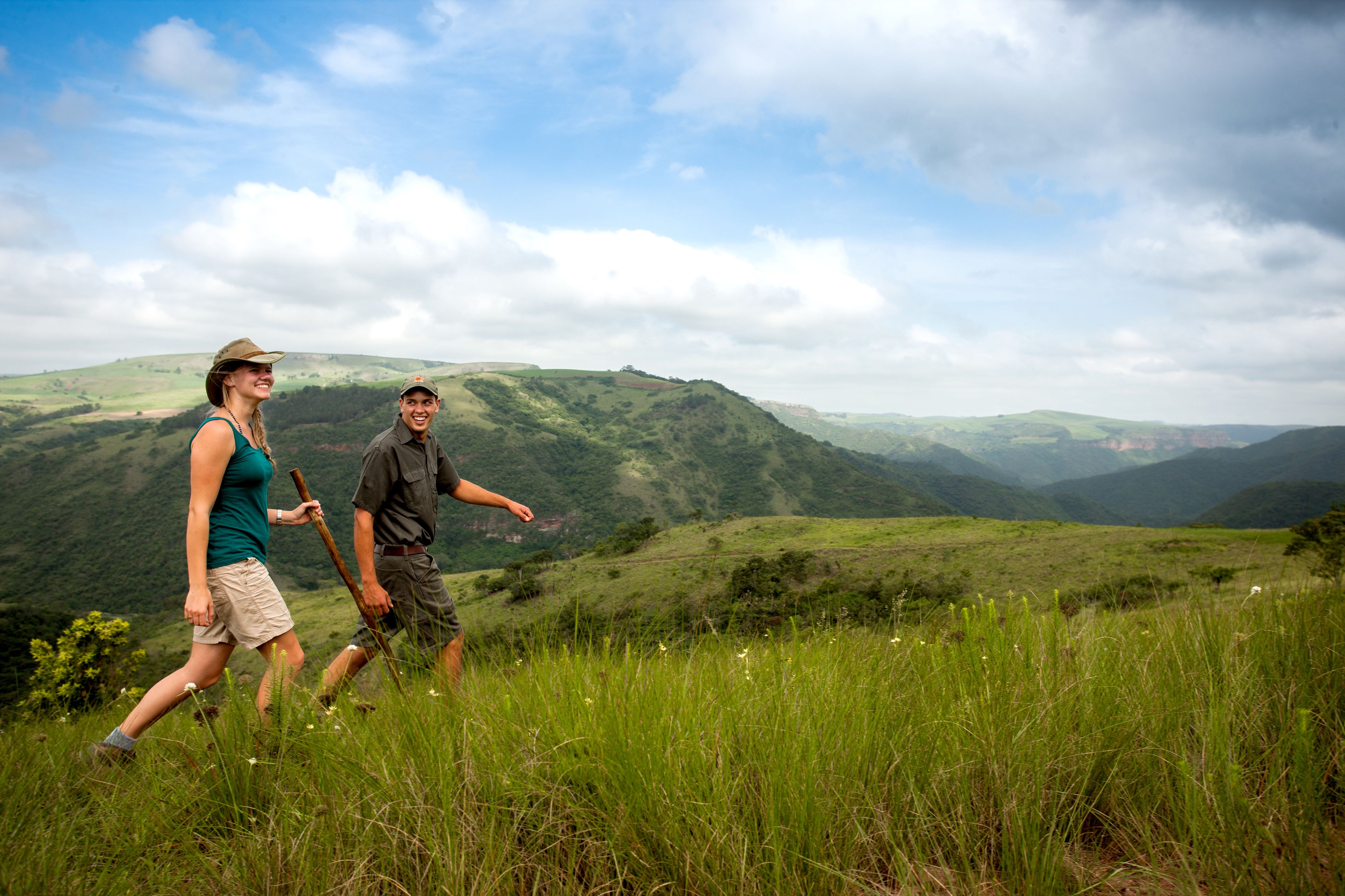 Two people walking on a grassy hill with a mountainous landscape in the background.