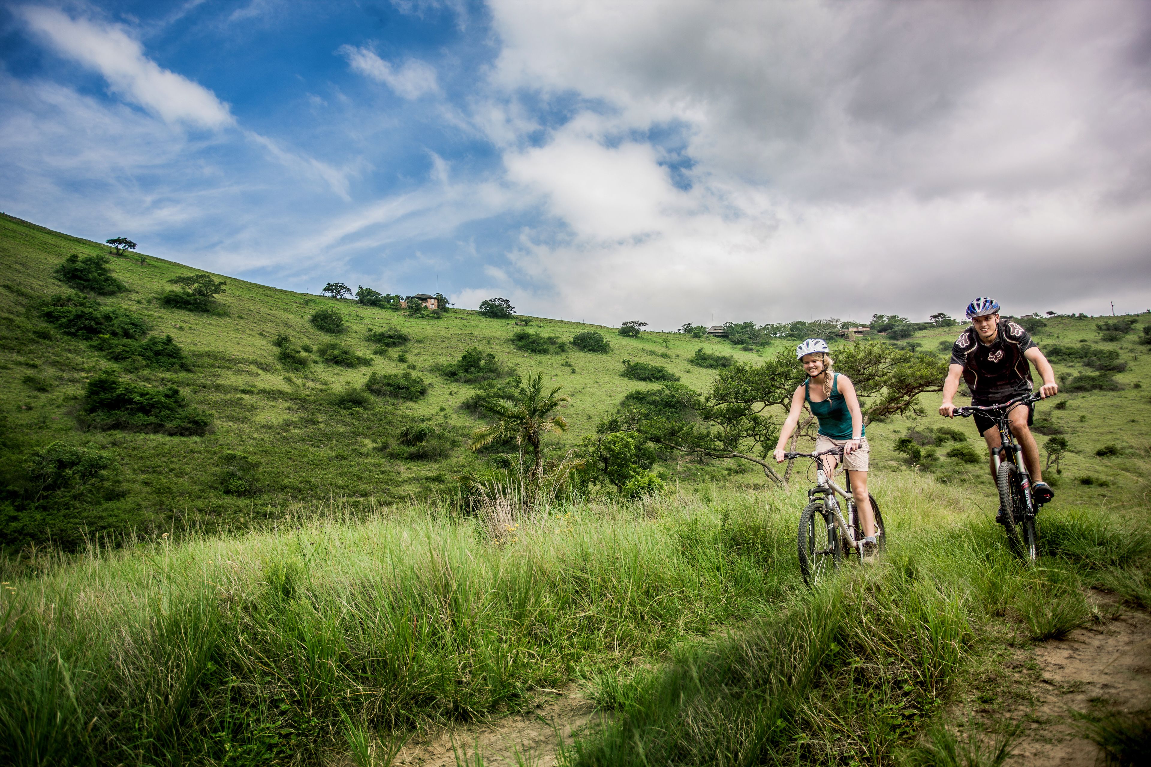 Two people riding bicycles on a grassy hill with a clear sky.