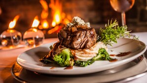 Une assiette de nourriture comprenant un steak, des brocolis et une garniture, le tout présenté sur un fond où l’on aperçoit une cheminée allumée.