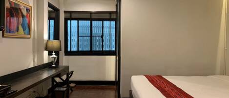 A hotel room with a bed, a desk, a chair, a window with blinds, and an air conditioning unit.