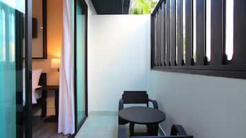 A small balcony with a table and chairs, a sliding glass door leading to a room with a bed and a nightstand, and a view of greenery outside.