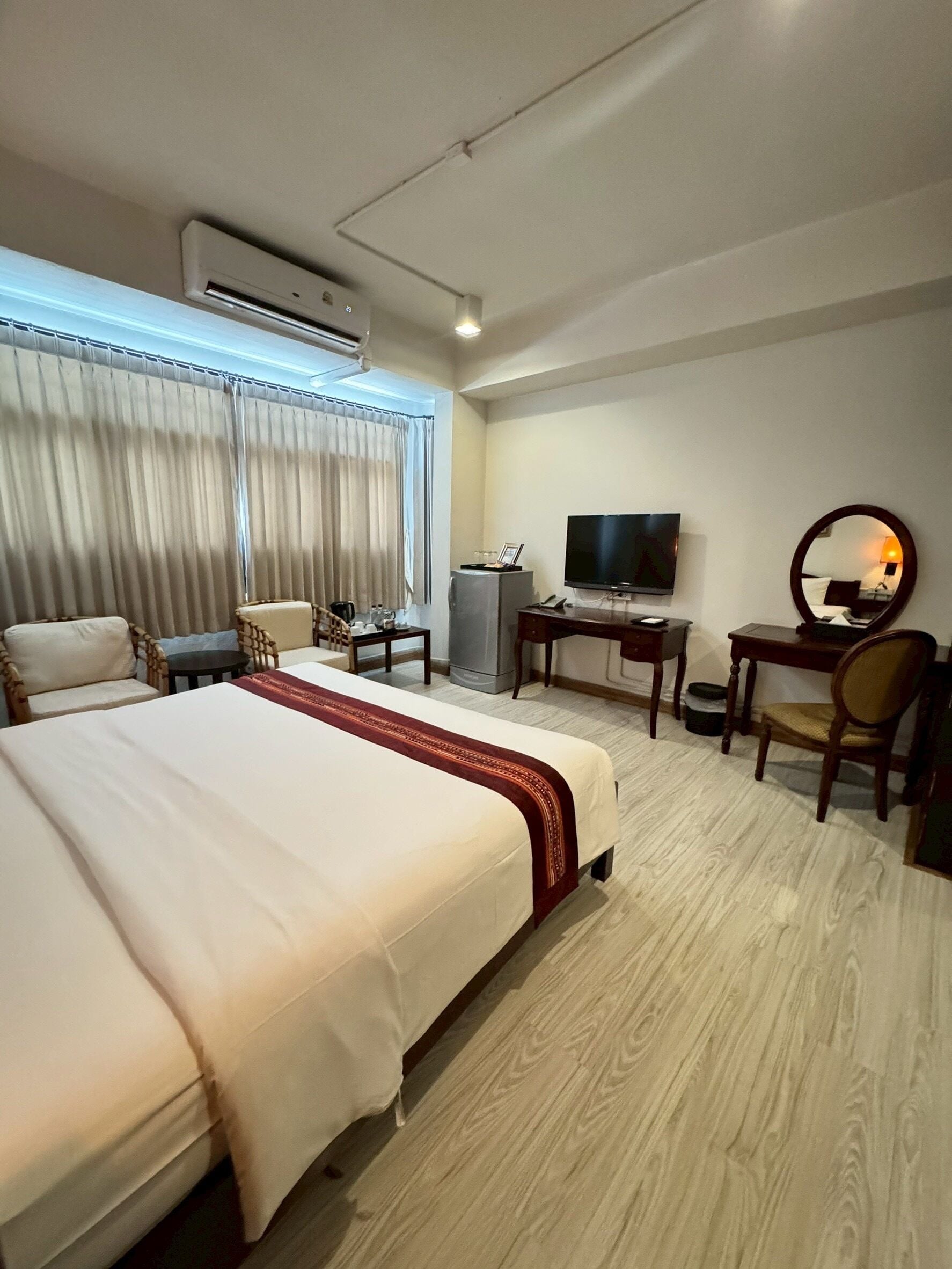 A hotel room with a large bed, a desk with a TV, a chair, a mirror, and an air conditioning unit.