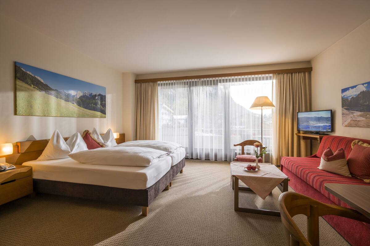 A hotel room with a bed, a sofa, a TV, and a painting of a mountain landscape.