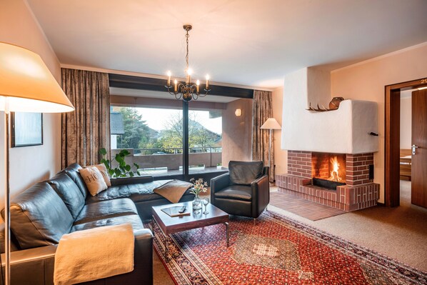 A living room with a leather sofa, a black armchair, a fireplace, and a large window with curtains. - Aparthotel Andreas Hofer (Kufstein)