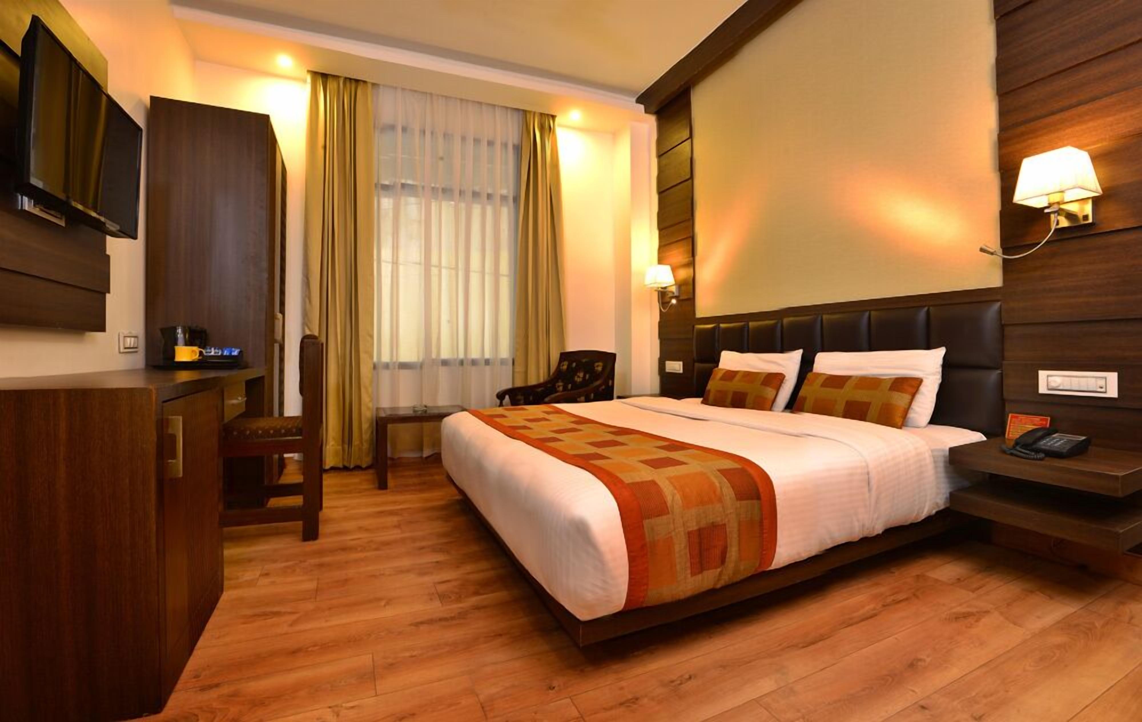 A hotel room with a large bed, wooden flooring, a desk with a chair, a television, and a wall-mounted lamp.