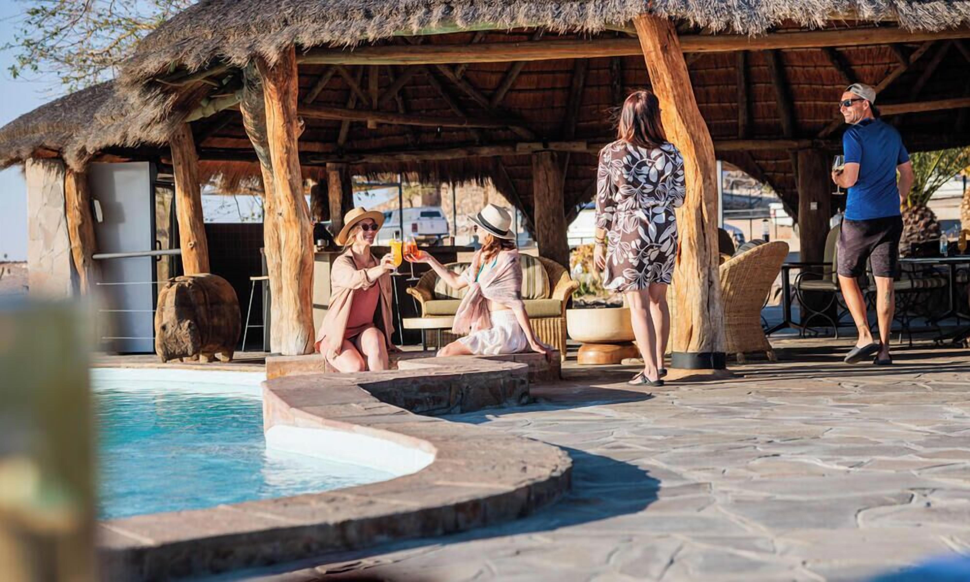 Un área de piscina con una estructura de techo de paja, suelo de piedra y una zona de estar con cojines.