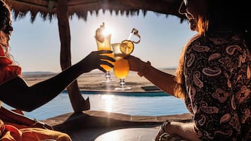 Two people toasting with drinks at a table with a view of a body of water and a distant landscape.