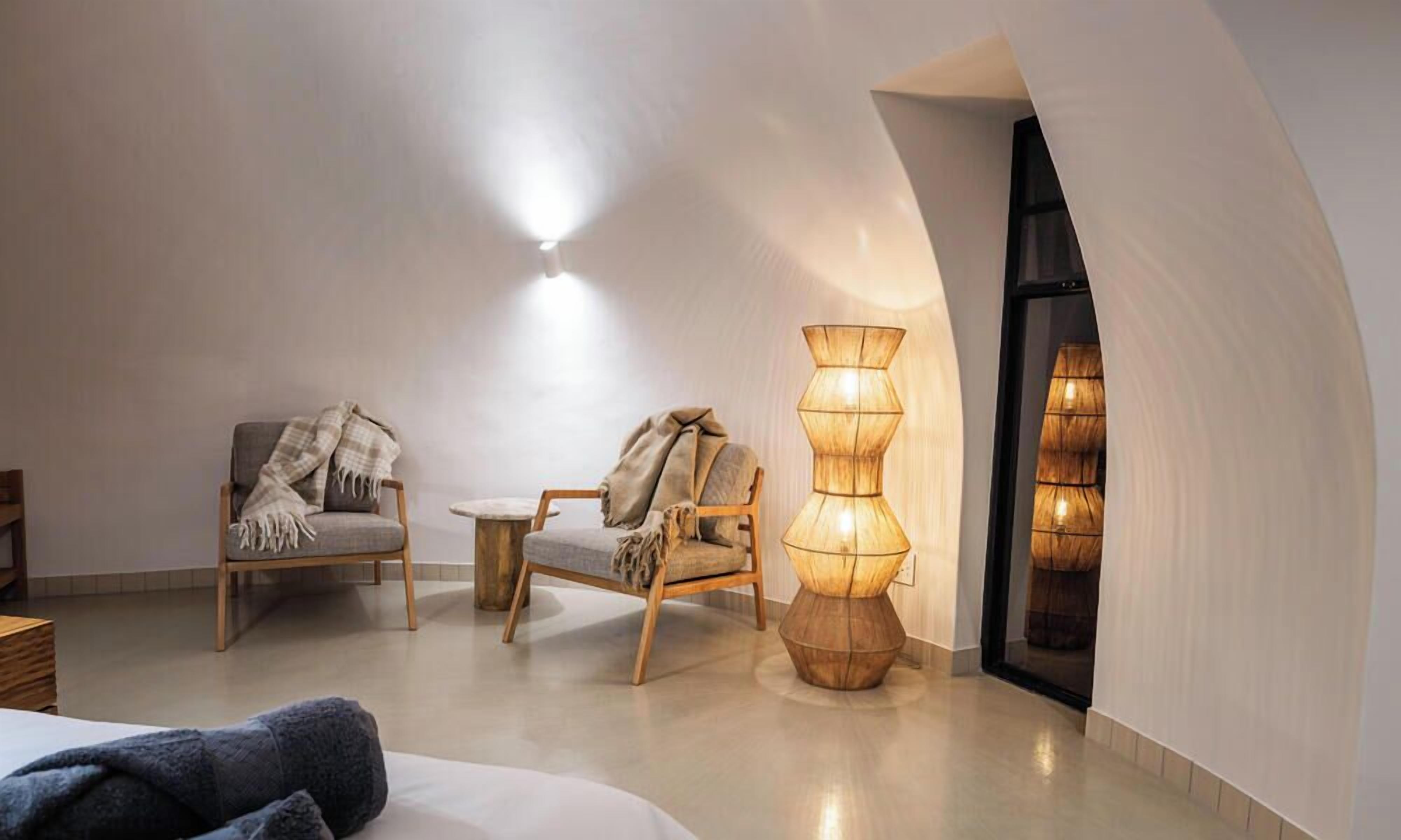 A modern interior with a high ceiling, a round wooden table, two chairs with blankets, and a tall, textured floor lamp.