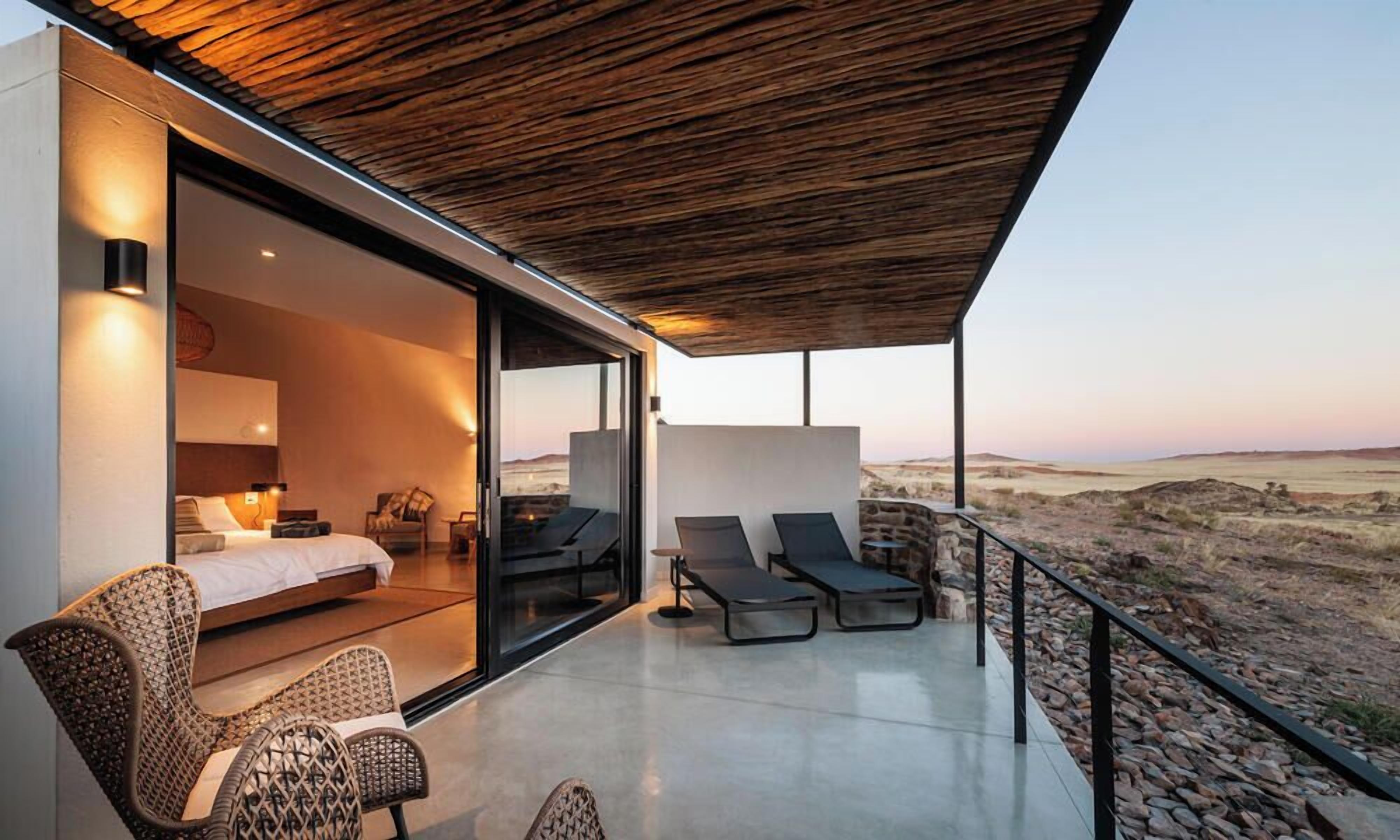 A modern bedroom with a glass door leading to a balcony, furnished with a bed, chairs, and a table.