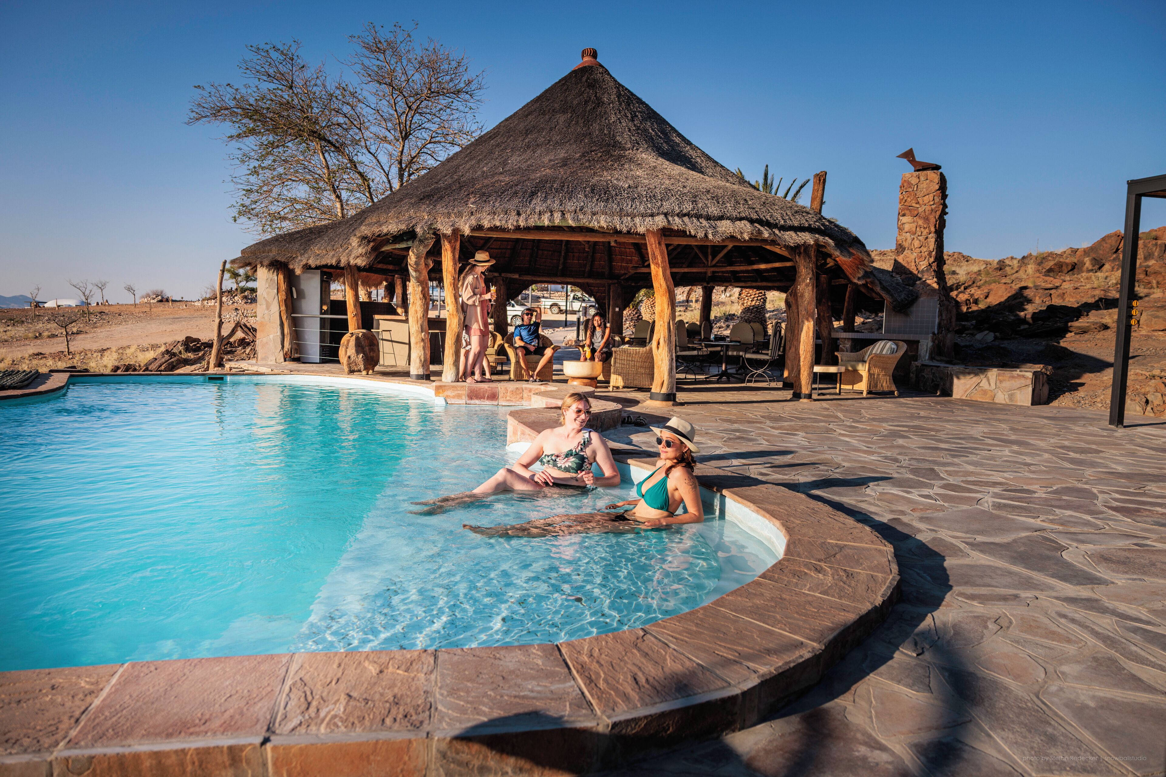 A pool area with a thatched-roof structure, stone flooring, and a clear blue pool.
