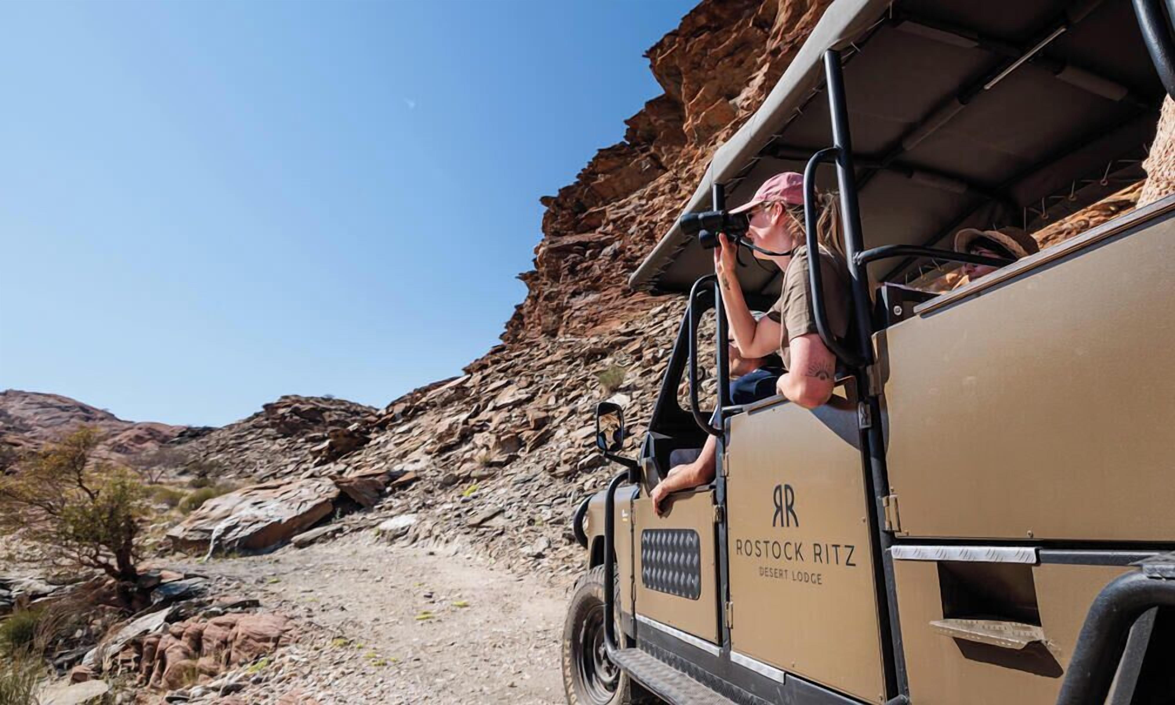 A desert lodge vehicle with a person hanging out the side, rocky terrain, and sparse vegetation.