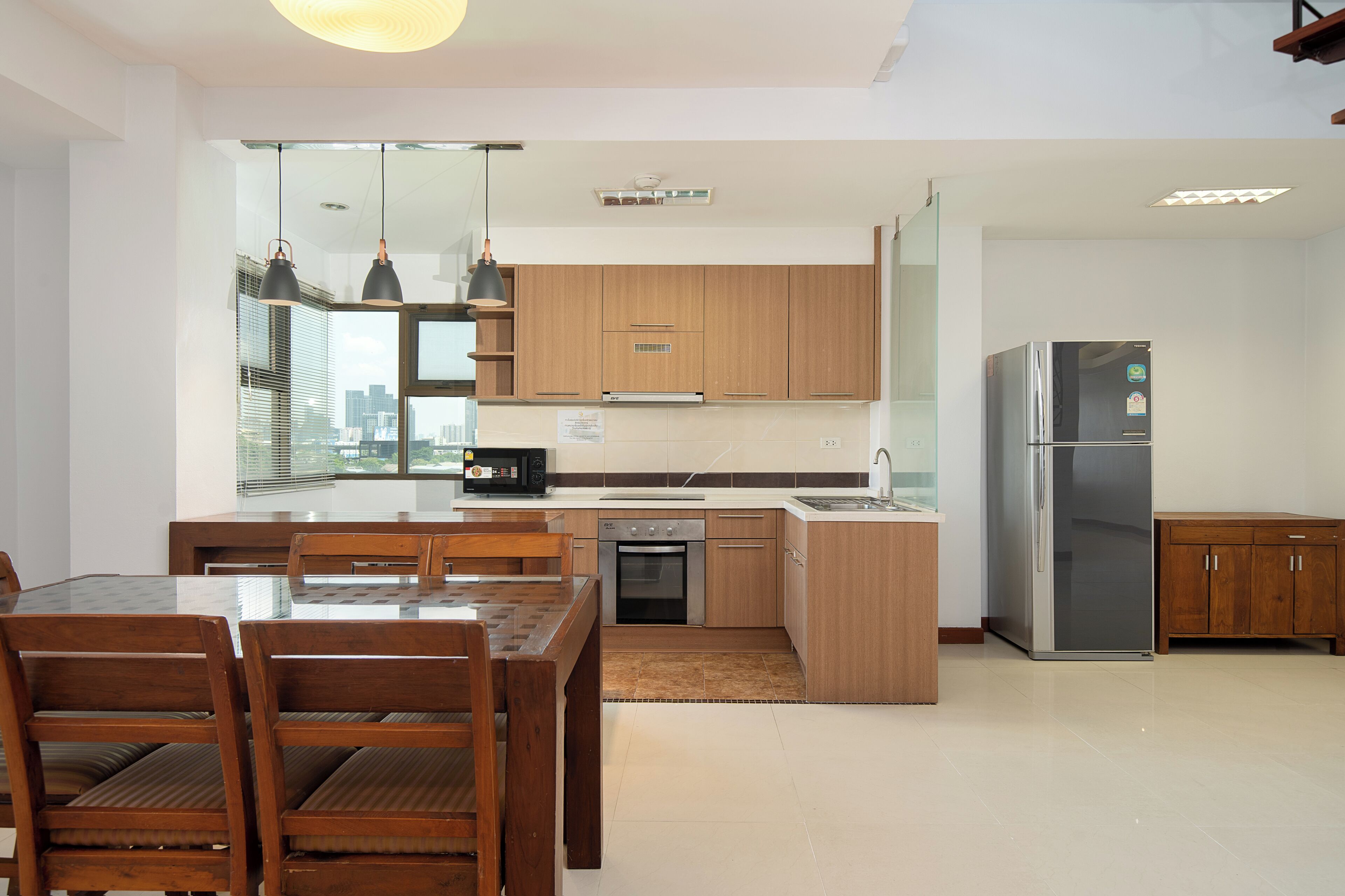 A modern kitchen with wooden cabinets, a stainless steel refrigerator, a built-in oven, and a dining area with wooden chairs and a glass table.