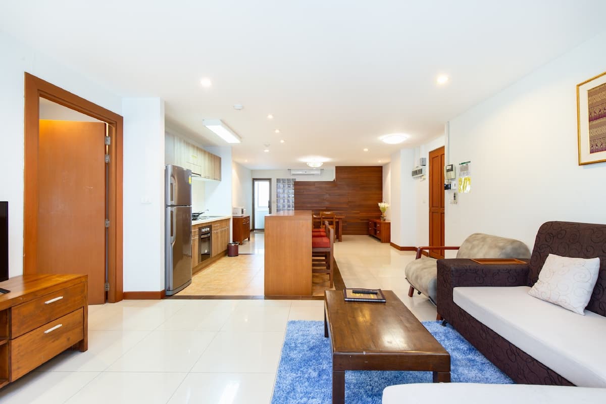 Una sala de estar moderna con sofá, mesa de centro y mueble para la televisión. Se observa un área de cocina con refrigerador y armarios. El piso es de madera y tiene un tapete azul.