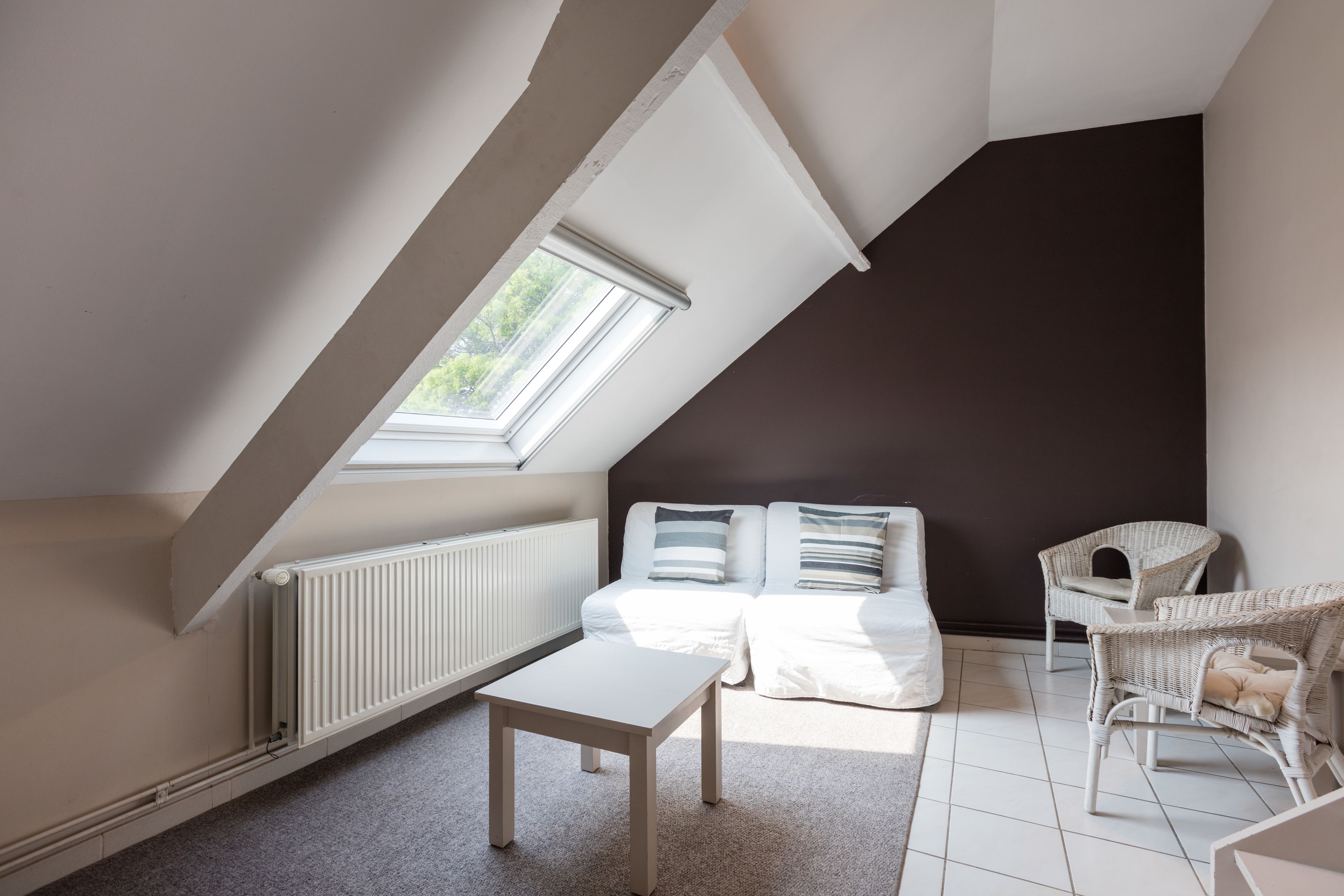 Een kamer met een schuine kap, een raam, een radiator, een witte tafel, een bank met kussens en rieten stoelen.