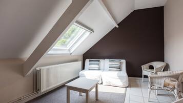 Een kamer met een schuine kap, een raam, een radiator, een witte tafel, een bank met kussens en rieten stoelen.