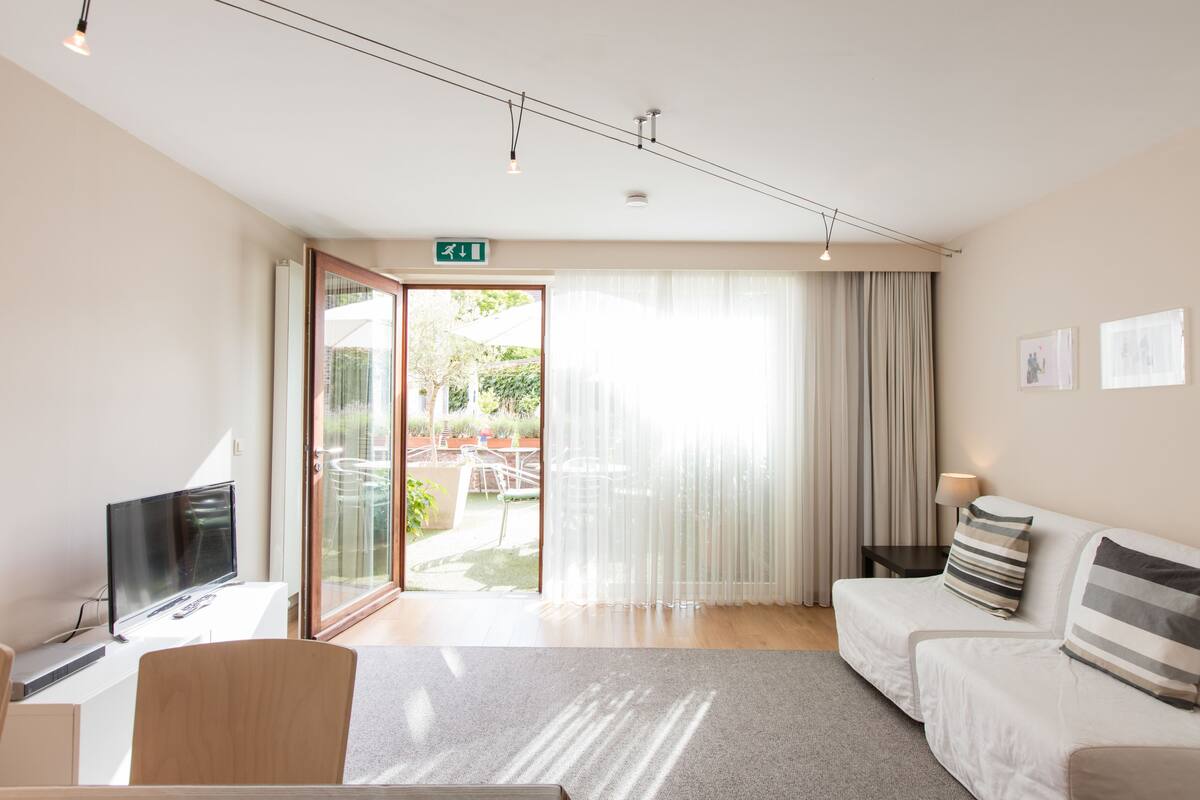 A modern hotel room with a flat-screen TV, a sofa, a small table, and a sliding glass door leading to a balcony.