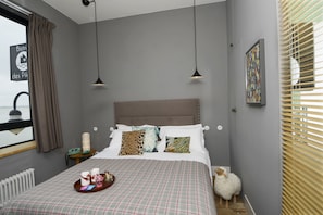 A bedroom with a bed, bedside table, lamp, and a window with blinds. - Hôtel Les Pilotes (Saint-Valery-sur-Somme)