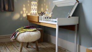 A room with a wooden floor, a white stool with a colorful blanket, a mirror on a white stand, and a shelf with various items.