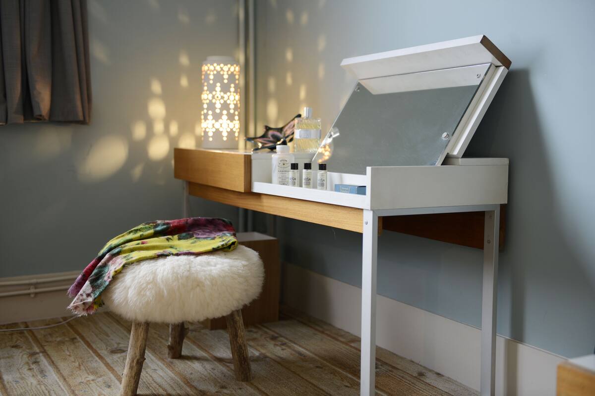 Een kamer met een houten vloer, een witte kruk met een kleurrijke deken, een spiegel op een witte standaard en een plank met diverse voorwerpen.