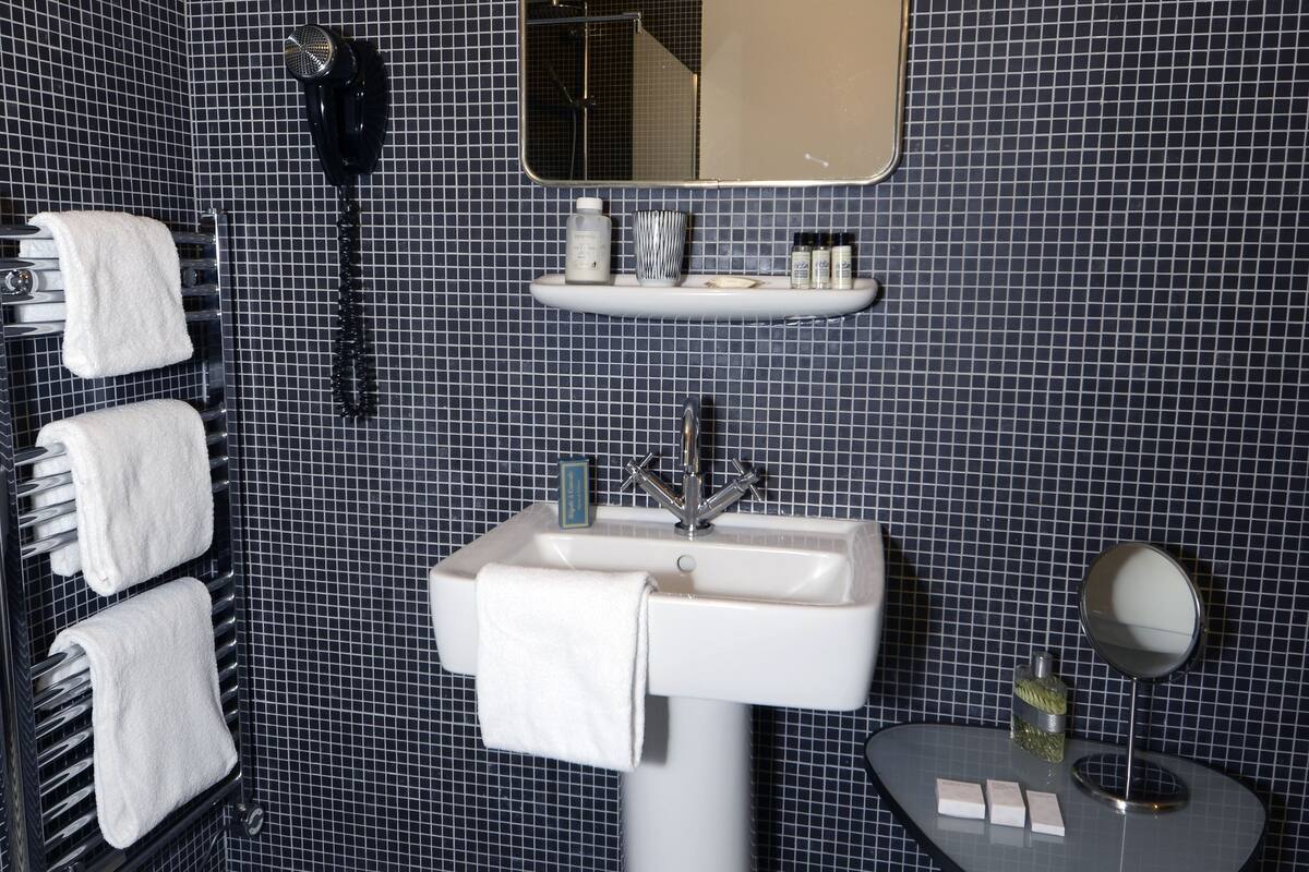 Een badkamer met een zwart-wit betegelde vloer, een wastafel met spiegel, een handdoekrek met witte handdoeken, een telefoon en een klein tafeltje met toiletartikelen.