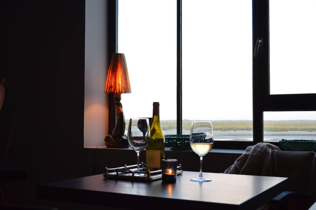 Een tafel met een fles wijn, twee glazen, een kaars en een lamp bij een raam met uitzicht.