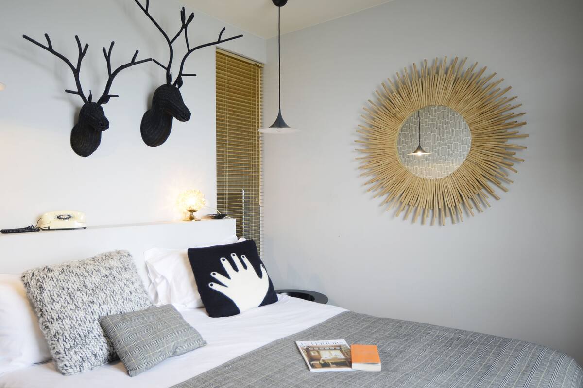 Een netjes opgemaakt slaapkamer met een bed, kussens, een boek, een spiegel aan de muur en een decoratief object.