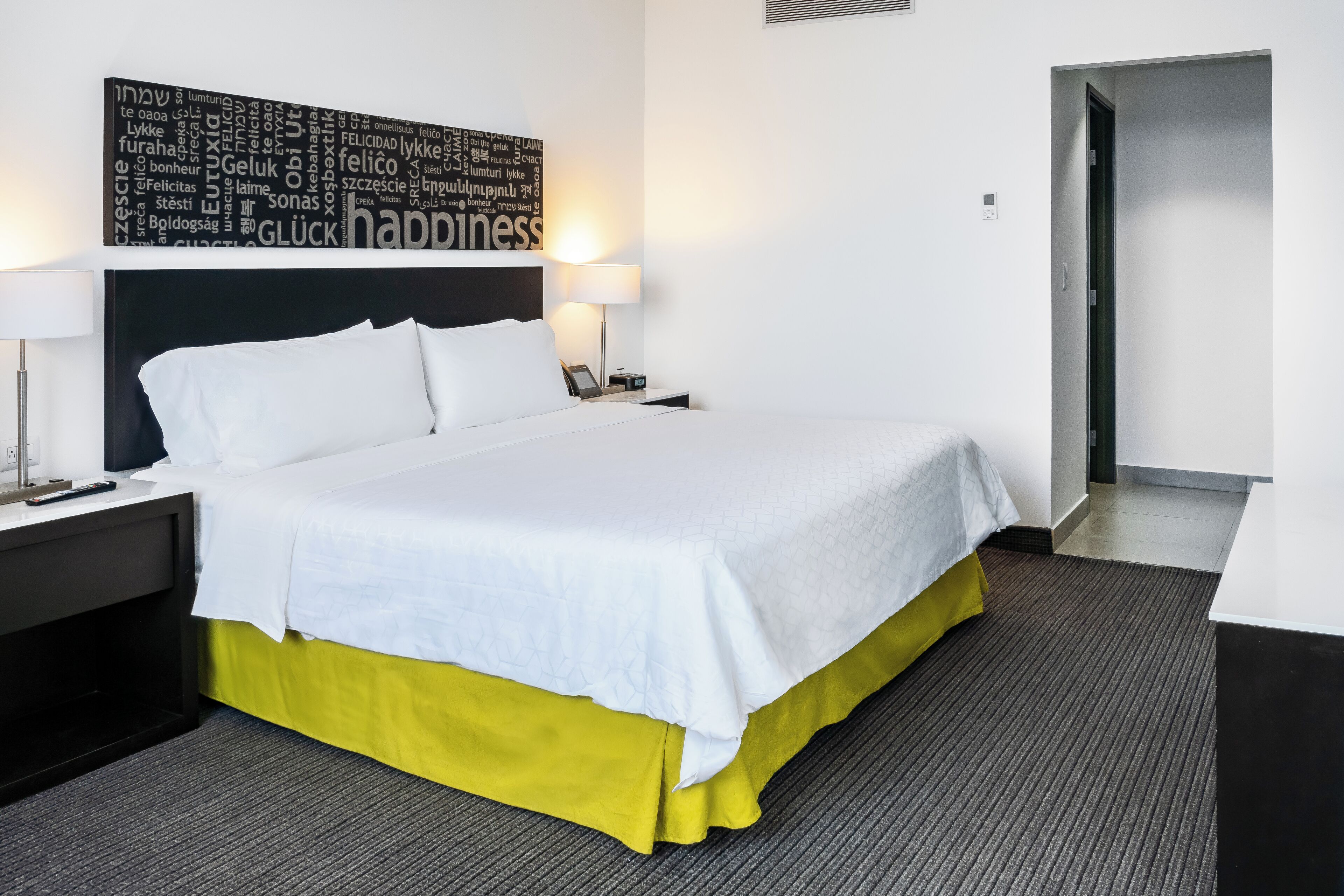 Una cama cuidadosamente hecha con sábanas blancas, una falda amarilla y un cabecero con diseño de nube de palabras. Hay dos mesitas de noche con lámparas, una alfombra gris y una puerta que da a otra habitación.