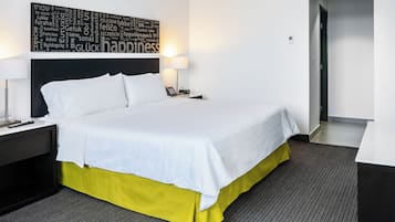 Una cama cuidadosamente hecha con sábanas blancas, una falda amarilla y un cabecero con diseño de nube de palabras. Hay dos mesitas de noche con lámparas, una alfombra gris y una puerta que da a otra habitación.