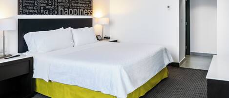 Una cama cuidadosamente hecha con sábanas blancas, una falda amarilla y un cabecero con diseño de nube de palabras. Hay dos mesitas de noche con lámparas, una alfombra gris y una puerta que da a otra habitación.
