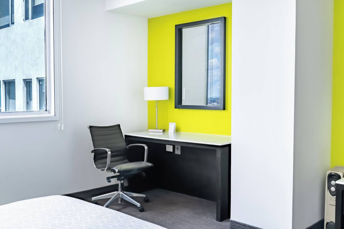 A hotel room with a bed, a desk with a chair, a window, and a bright yellow accent wall.