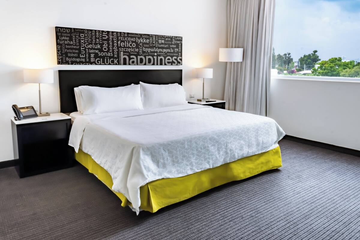 A hotel room with a large bed, two bedside tables, a wall-mounted word cloud, and a window with a view.