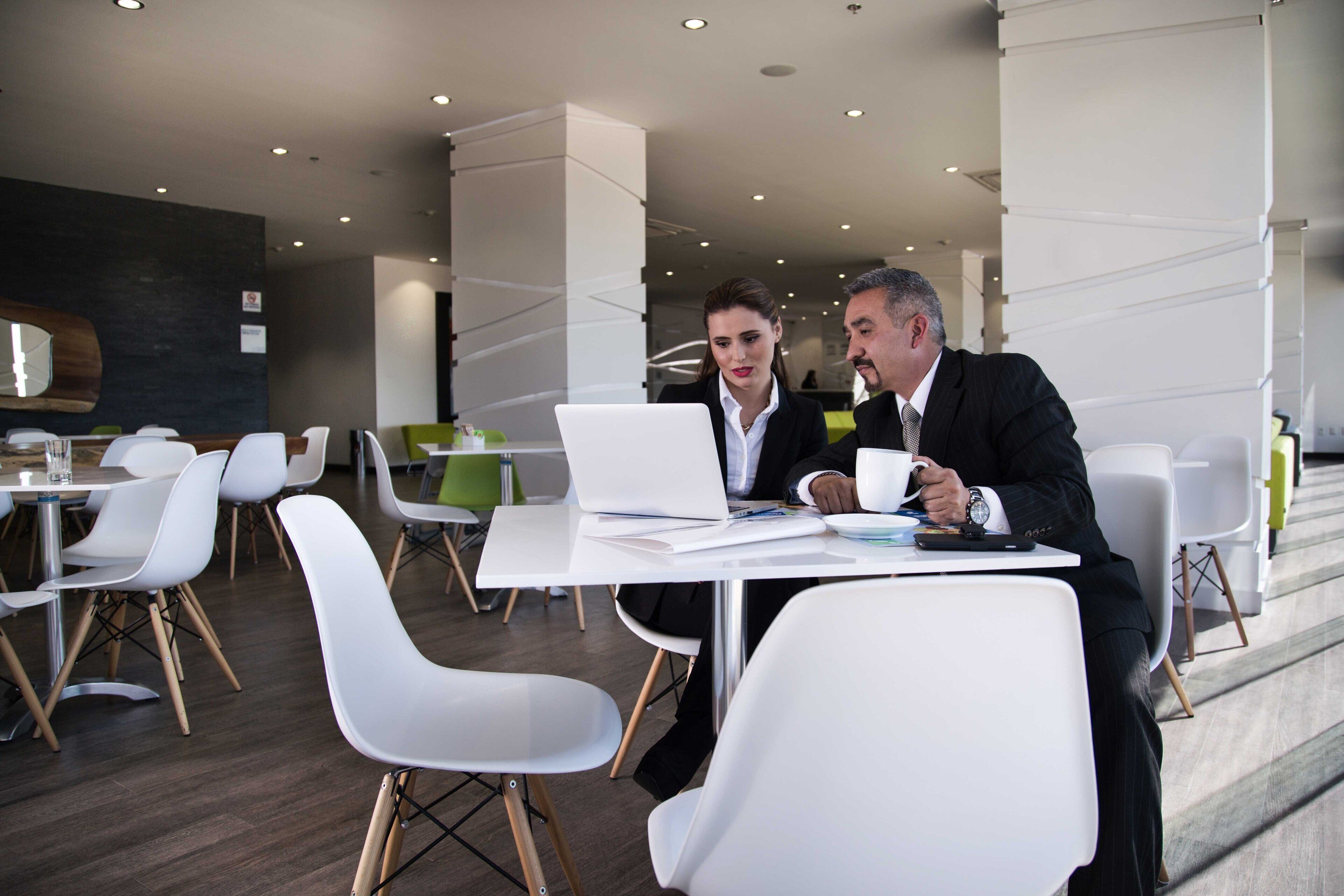 Un espacio interior moderno con mobiliario blanco, incluyendo mesas y sillas, y una persona usando una computadora portátil.