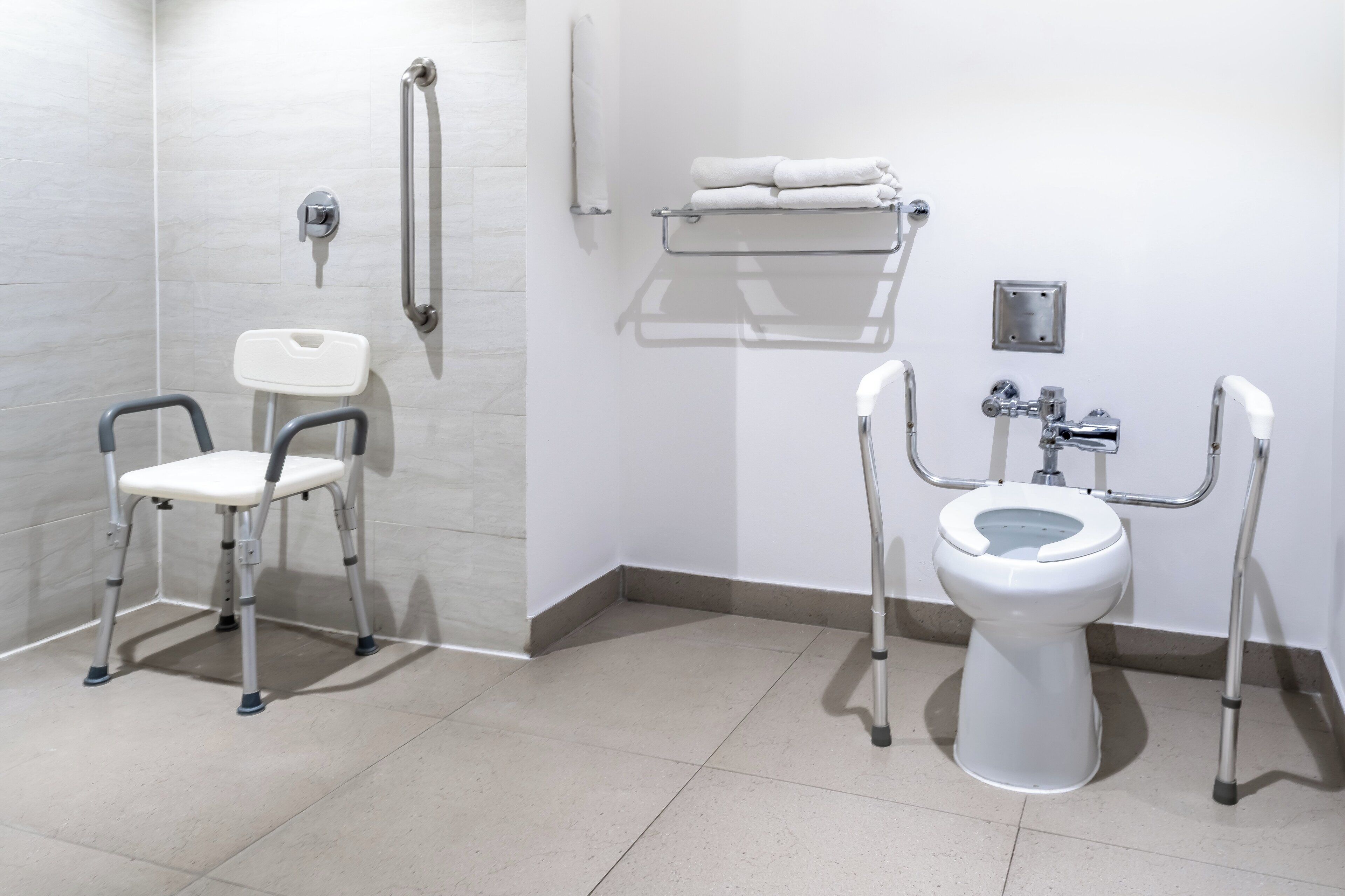 Een badkamer met een wit toilet, een witte wasbak en een witte douche. Er zijn handgrepen naast het toilet en een witte stoel.
