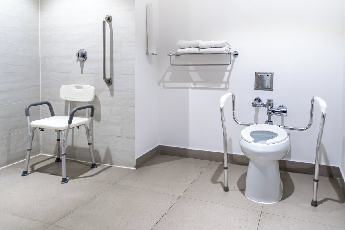 A bathroom with a white toilet, a white sink, and a white shower. There are grab bars next to the toilet and a white chair.