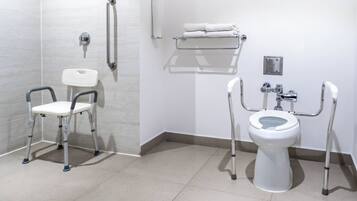 A bathroom with a white toilet, a white sink, and a white shower. There are grab bars next to the toilet and a white chair.