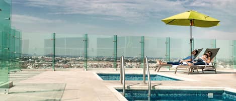A rooftop pool area with glass railings, a pool, and a view of a cityscape.