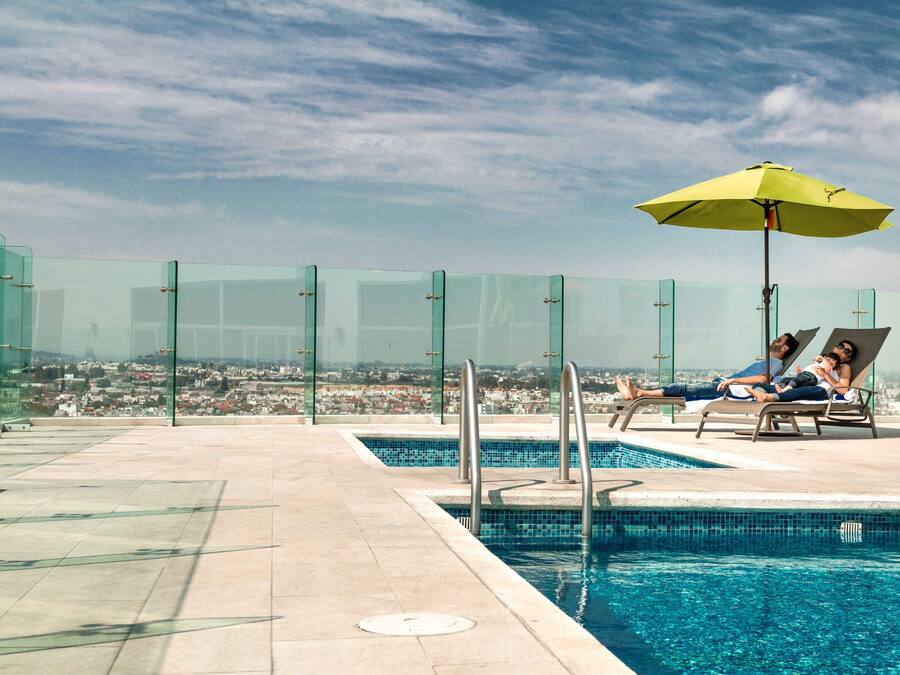 A rooftop pool area with glass railings, a pool, and a view of a cityscape.